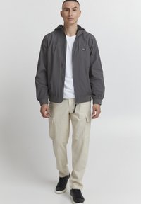 Giacca leggera grigia con cappuccio, chiusura con zip, polsini elastici, abbinata a pantaloni cargo beige e sneakers nere. Design e materiali semplici.