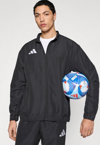 Μαύρο αθλητικό μπουφάν με φερμουάρ μπροστά, ελαφρύ ύφασμα, με λευκά διακριτικά λογότυπα Adidas και ελαστικά μανίκια, κρατημένο με πολύχρωμη μπάλα ποδοσφαίρου.
