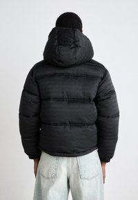 Black quilted puffer jacket with a hood, featuring a subtle repeating logo pattern. Made from a smooth, glossy material.