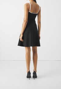 Schwarzes ärmelloses Kleid mit tailliertem Oberteil und ausgestelltem Rock, ausgestattet mit weißen Gurtakzenten und einer glatten Stoffstruktur. Tragen mit schwarzen hohen Schuhen.
