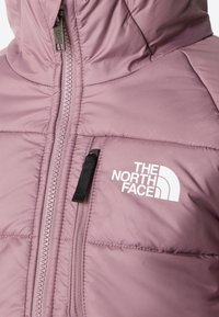 Zbliżenie na fioletową puchową kurtkę z zamkiem błyskawicznym z przodu i kieszenią na piersi, z białym logo The North Face na klatce piersiowej.