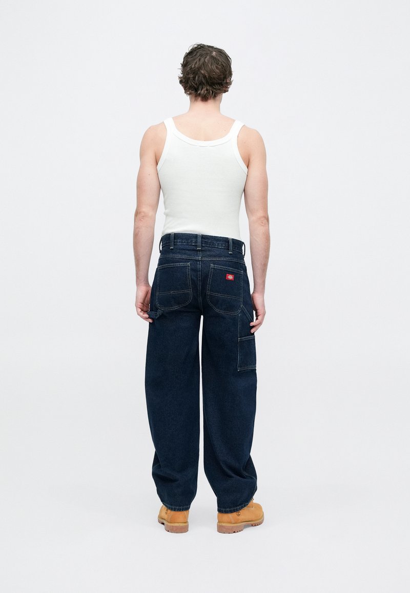 Pantalon cargo en denim bleu foncé avec plusieurs poches, une coupe ample et un logo en patch à l'arrière ; associé à un débardeur blanc et des chaussures couleur beige.