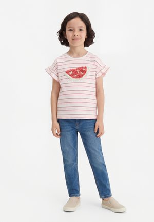 Junges Mädchen steht, trägt ein gestreiftes pinkes T-Shirt mit strukturiertem Wassermelonen-Design, blaue Jeans und beige Slipper, neutraler Hintergrund.
