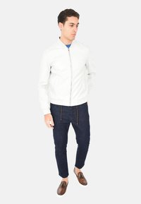 Giacca bomber bianca in pelle con zip frontale, abbinata a pantaloni in denim scuro con un cordoncino intrecciato, e mocassini marroni con suola bianca.