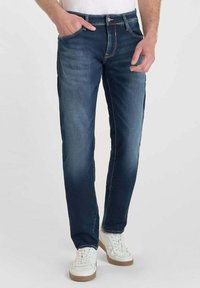 Vaqueros de mezclilla azul oscuro con un corte recto, que cuentan con cinco bolsillos y costuras doradas. Combinados con zapatillas blancas para un contraste.