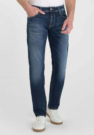 Vaqueros de mezclilla azul oscuro con un corte recto, que cuentan con cinco bolsillos y costuras doradas. Combinados con zapatillas blancas para un contraste.