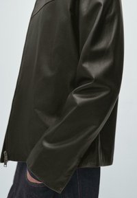 Svart läderjacka med en slät yta, dragkedjestängning och en avslappnad passform. Ärmarna sträcker sig till mitten av underarmen, vilket visar en ren design.