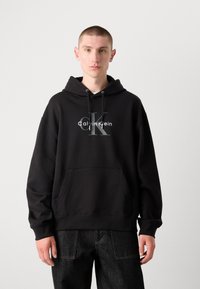 Crna dukserica s kapuljačom i prednjim džepom, s velikim sivim logotipom koji glasi "Calvin Klein" i istaknutim slovom "K."