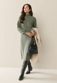 Abito in maglia verde oliva con collo alto, dettagli a coste sulle maniche e bottoni decorativi dorati. Abbinato a stivali neri al ginocchio e borsa nera.