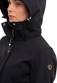 Veste en softshell noire avec un col montant et une capuche amovible ; dotée de quincaillerie argentée, d'une fermeture éclair et d'un patch logo sur l'épaule gauche.