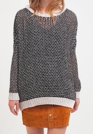 Femme portant un pull en maille noir et blanc avec un motif géométrique sur une jupe en daim beige à boutons devant.