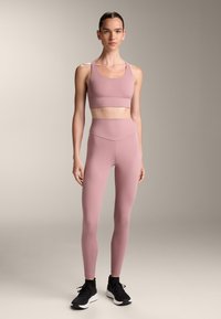 Világos rózsaszín sportmelltartó és hozzá illő, magas derekú leggings, rugalmas anyagból, fekete sportcipővel kontrasztban.