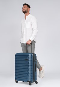 Valigia rigida blu con scanalature testurizzate, ruote doppie e manico telescopico, abbinata a vestiti leggeri e sneaker.