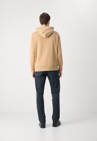 Felpa con cappuccio color marrone chiaro, maniche lunghe, polsini e orlo a costine, abbinata a jeans blu scuro e scarpe nere, vista da dietro.