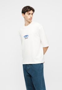 Jeune homme portant un T-shirt blanc oversize avec un petit logo bleu, associé à un jean bleu ample, regardant sur le côté avec les mains dans le dos.