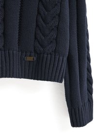 Mörkblå stickad tröja med kabelmönster, ribbade ärmslut och nederkant. Liten bronsskylt med "POLO CLUB" som detalj nära nederkanten.