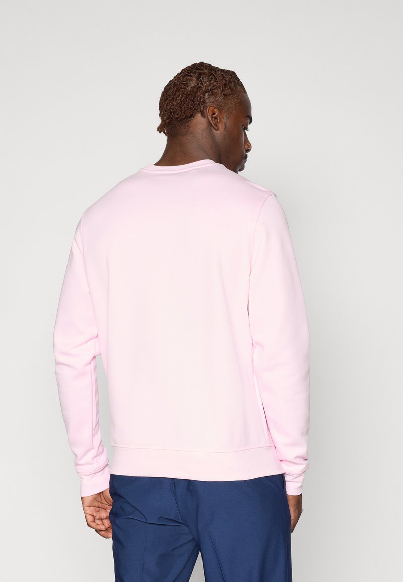 Homme avec des cheveux tressés portant une chemise rose clair à manches longues et un pantalon bleu marine, de dos contre un fond clair uni.