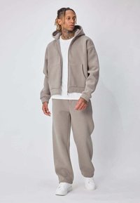 Homme tatoué portant un sweat à capuche beige zippé, un pantalon assorti, un t-shirt blanc et des baskets blanches, debout devant un fond uni.