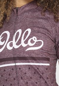 Mörkröd cykeltröja med en vit "Hello"-logotyp, halvdragkedja fram och prickmönster med horisontella randiga detaljer.