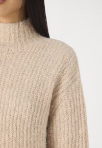 Beżowy sweter dziany z wysokim kołnierzem, wykonany z miękkiej, teksturowanej tkaniny i z prążkowanym wzorem. Zbliżenie na obszar ramienia.