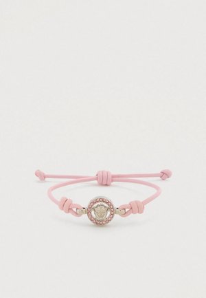 Rozā ādas rokassprādze ar apaļu metāla piekariņu, kurā attēlots lauva galva un malas rotātas ar rozā rhinestone akmentiņiem. Regulējams aizdare ar sasienamu lenti.