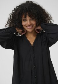 Black blouse with a V-neckline, intricate lace trim, and decorative buttons. Features long, gathered sleeves and a soft, textured fabric.