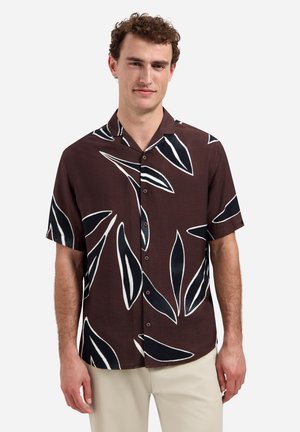 Mann trägt ein braunes kurzärmeliges Hemd mit großen schwarz-weißen Blattmustern, kombiniert mit beigen Hosen, stehend.