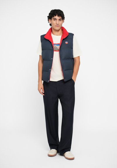 Navy puffer vest with a red inner collar, white t-shirt featuring a mountain graphic, striped black trousers, and light shoes.