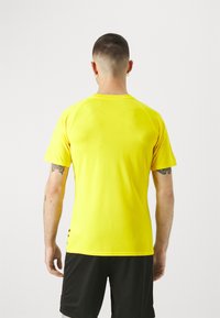 Camiseta deportiva amarilla con textura transpirable, mangas cortas y escote redondeado, combinada con pantalones cortos negros. Vista trasera mostrada.