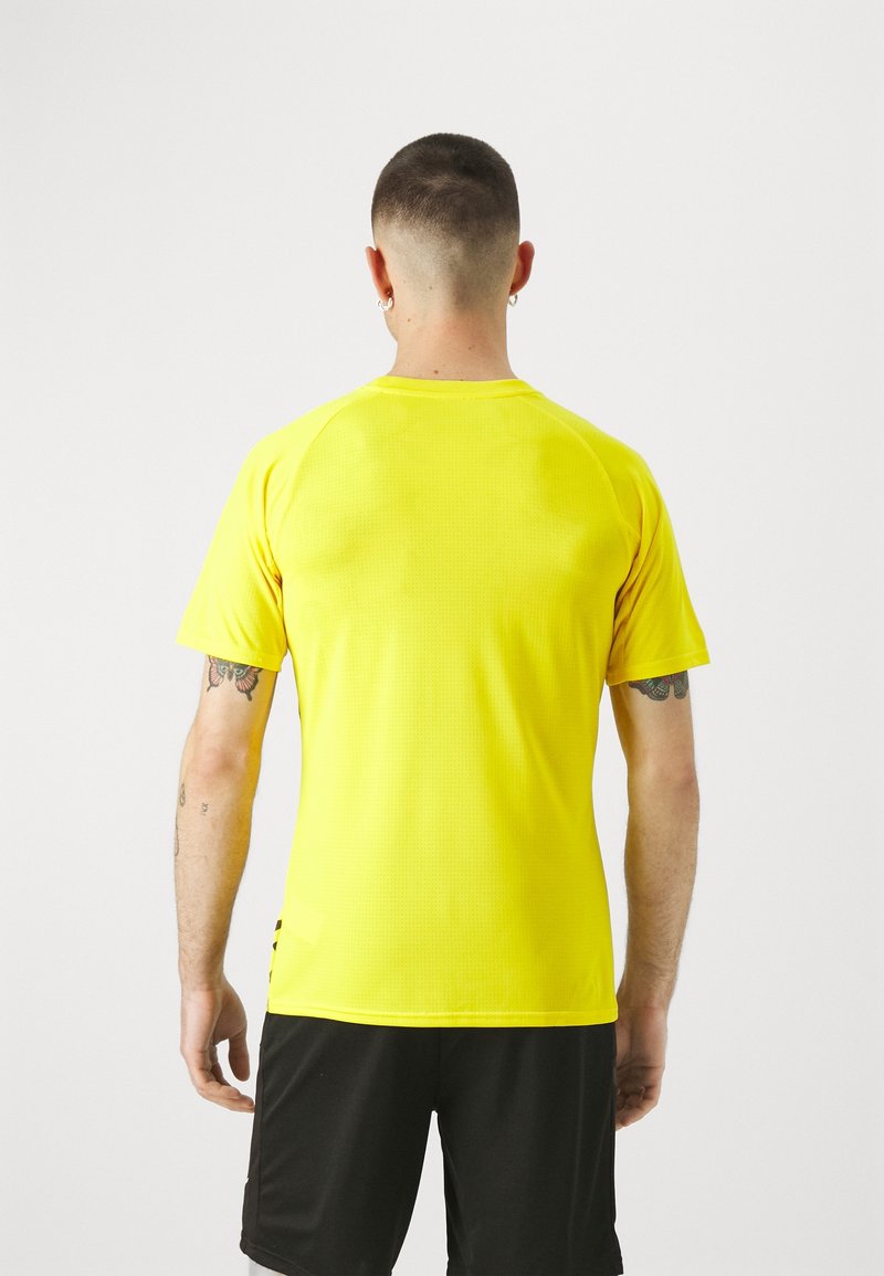 Camiseta deportiva amarilla con textura transpirable, mangas cortas y escote redondeado, combinada con pantalones cortos negros. Vista trasera mostrada.