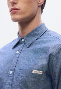 Svetlo modra chambray srajca z ovratnikom na gumbe, z levim žepom na prsih in majhno etiketo "Calvin Klein". Gladek tekstura, prilagojen kroj.