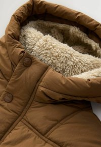 Veste matelassée marron avec doublure polaire couleur crème visible à l'intérieur de la capuche et deux boutons marron au niveau du col.