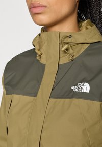 Φόρεμα σε ελιά πράσινο με σκουρό πράσινο ανώτερο πάνελ, που διαθέτει ψηλό γιακά, κορδόνια με ρυθμιστικά και λευκό λογότυπο στο στήθος.