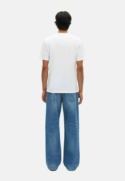 Uomo visto di spalle che indossa una maglietta bianca a maniche corte e jeans in denim blu larghi su uno sfondo semplice.