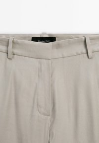 Pantalon beige confectionné sur mesure avec passants pour ceinture et fermeture éclair avant dissimulée, doté d’une étiquette noire portant la mention « Massimo Dutti » à l’intérieur de la ceinture.