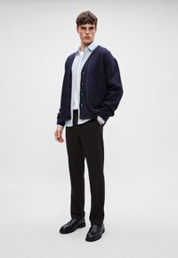 Sötétkék kötött kardigán egy világoskék csíkos ing fölött, fekete nadrággal és cipővel párosítva. Gombokkal és bordázott mandzsettákkal rendelkezik.