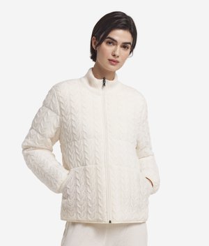 Femme portant une veste blanche en maille torsadée zippée avec des poches, debout devant un fond clair uni, mains dans les poches.