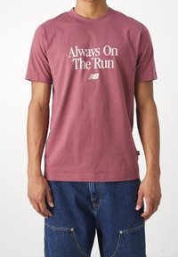 Man in een dusty rose t-shirt met de tekst "Always On The Run" en het New Balance logo, gecombineerd met blauwe denim broeken met voorzakken.