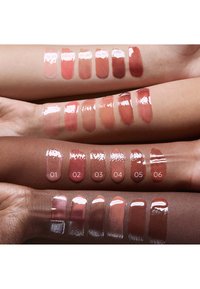 Échantillons de rouge à lèvres brillant dans six teintes nude appliqués sur quatre bras avec des carnations variées, montrant la diversité des couleurs et la finition.