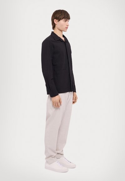 Black, textured long-sleeve shirt with a collar, paired with light gray trousers and white sneakers, presented in a side view.
