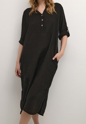 Kvinde iført en løs sort midi-kjole med tre knapper på knaplisten og oprullede ærmer, den ene hånd i lommen, stående mod en ensfarvet væg.