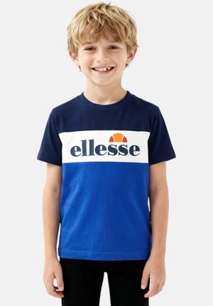 Garçon blond souriant portant un T-shirt bicolore bleu et marine avec le logo "ellesse", debout devant un fond clair uni.