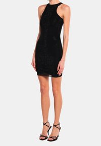 Robe noire sans manches mini avec un design ajusté, présentant une surface texturée de petits clous. Associée à des talons hauts noirs à lanières.
