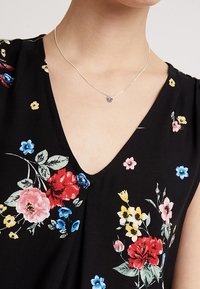 Frau in einem schwarzen V-Ausschnitt-Oberteil mit mehrfarbigem Blumenmuster und einer zarten Silberkette mit einem kleinen Herzanhänger.