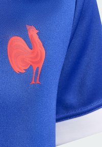 Plava majica s teksturiranim tkanjem. Crveni pijetao kao logo vidljivo prikazan na prsnom području. Bijeli naglasak na rubovima rukava.