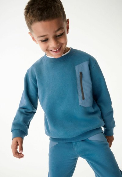 Felpa blu con tasca con zip, tessuto testurizzato e polsini a costine. Abbinata a pantaloni blu coordinati con accenti simili.