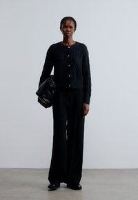 Μαύρη ζακέτα με πλεκτό σχέδιο και στρογγυλή λαιμόκοψη, συνδυασμένη με μαύρο παντελόνι με φαρδιά πόδια. Κρατά μια μεγάλη μαύρη τσάντα, σε απλό φόντο.