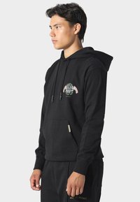 Giovane uomo in felpa nera con cappuccio e pantaloni abbinati con logo PAS DE MONACO, in piedi di lato contro uno sfondo chiaro e semplice.