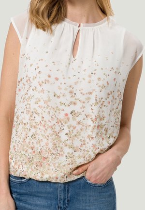 Mujer con blusa blanca sin mangas con pequeño estampado floral rosa y verde, combinada con jeans de mezclilla azul, con la mano en el bolsillo.