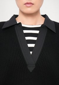 Schwarzes strukturiertes Top mit einem V-förmigen Kragen, das über einem schwarz-weiß gestreiften Hemd getragen wird. Der Kragen ist flach und spitz und hebt sich von den darunterliegenden Mustern ab.